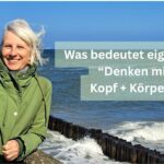 Birgit Krüger in grüner Jacke lächelt am windigen Meer; Text im Bild: „Was bedeutet eigentlich Denken mit Kopf + Körper?“