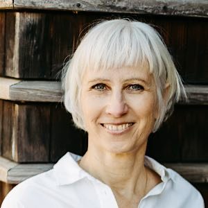 Porträt von Birgit Krüger vor einer Holzwand, weißes Haar, Blick in die Kamera